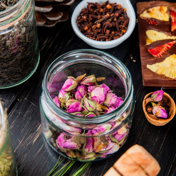 side view of rose buds in a glass jar, dry black tea leaves, clove spice and chocolate bar with fruits on black wooden background